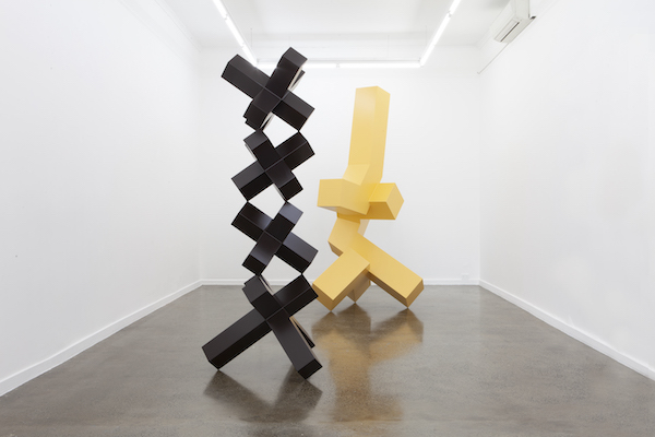 Two large-scale abstract sculptures of lines and forms stand In a galley space surrounded by white walls. The sculpture situates on the left is in chocolate black, and the one on the right is in butter yellow.