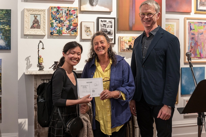 Three individuals stand together in an art gallery, with the
person on the left holding a certificate and all three smiling. Behind them is a wall filled with small, colourful artworks, creating a vibrant backdrop for what appears to be a celebratory moment.
