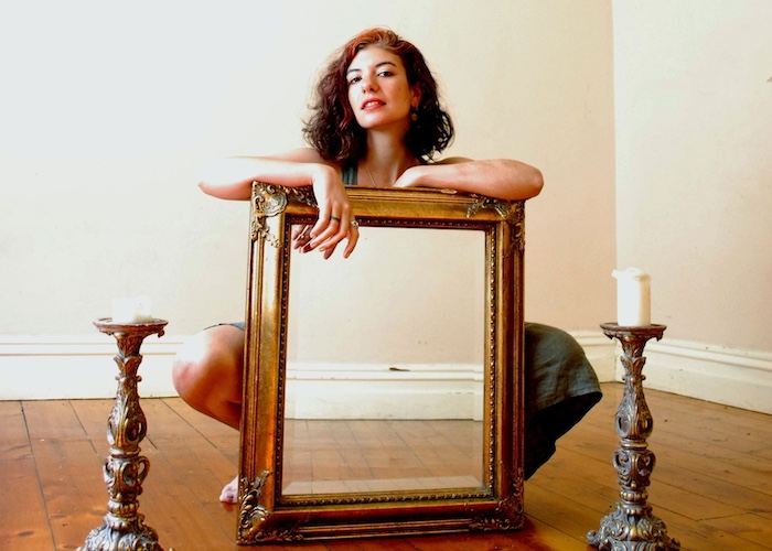 A woman kneels behind an ornate, empty gold frame placed on a polished wooden floor, flanked by two antique-style candlesticks. The natural light streaming in highlights the warm tones of the scene, creating an intimate and vintage-inspired atmosphere.