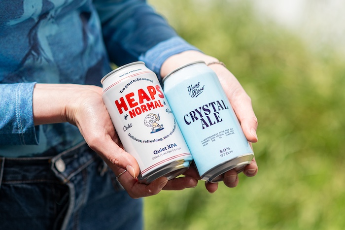 Close-up of a person's hands holding two cans of beers. The one on the left features red colour font 'Heaps Nomal', the one on the right is in light blue package with text 'Crystal Ale'.