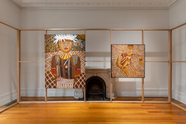 This image captures a contemporary art installation featuring two striking paintings. Both artworks are set within a minimal wooden frame structure, centered around a small, decorative fireplace in a gallery space with polished wooden floors. On the left, a vibrant portrait showcases a figure adorned with traditional patterns and a large feathered headdress, rendered in earth tones with bold white accents. On the right, a smaller piece features a similarly detailed design, with intricate geometric patterns and a crowned figure.