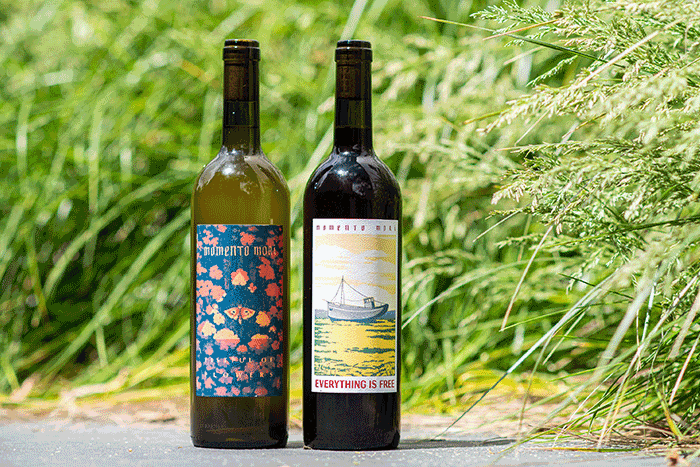 Two bottles of wine from the label "Momento Mori" stand side by side against a backdrop of lush green grass. Their artistic labels, one adorned with floral patterns and the other featuring a serene boat scene, suggest a thoughtful and creative winemaking approach.