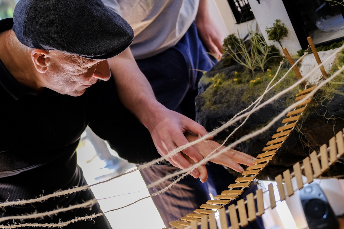 The image shows a close-up of two individuals engaged in a tactile exploration of an art installation. A person wearing a dark flat cap and a black sweater is leaning forward closely examining the installation while touching a miniature wooden bridge, which is suspended by strings. Another person's hand is gently guiding the first person's hand over the bridge.