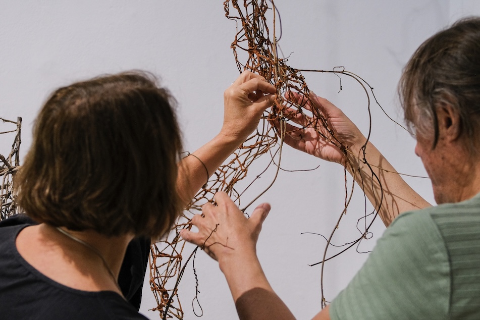 The image shows two individuals facing away from the camera, engaging with an intricate structure made of thin, intertwined branches. Their hands delicately manipulate the twigs. The branches form a complex web, displaying a mix of natural tones from dark brown to reddish hues. 