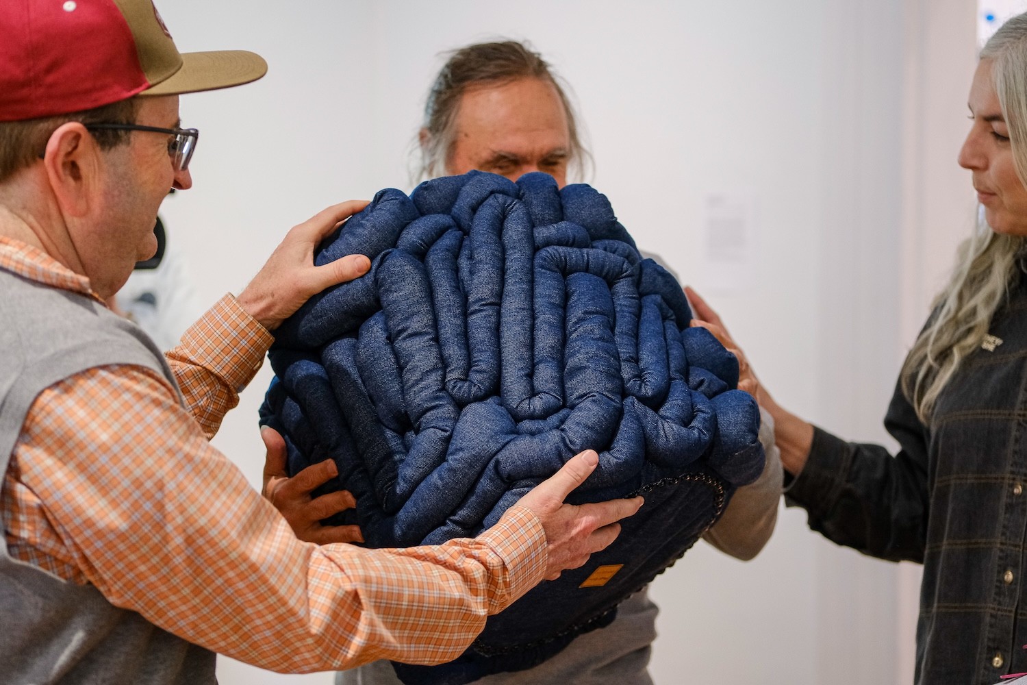 The image shows three individuals collectively holding a large, cushion-like object. The object is made of tightly packed, deep blue, quilted fabric and has a textured, soft appearance. The fabric is coiled and stitched in a manner that creates a series of winding, rounded shapes. 