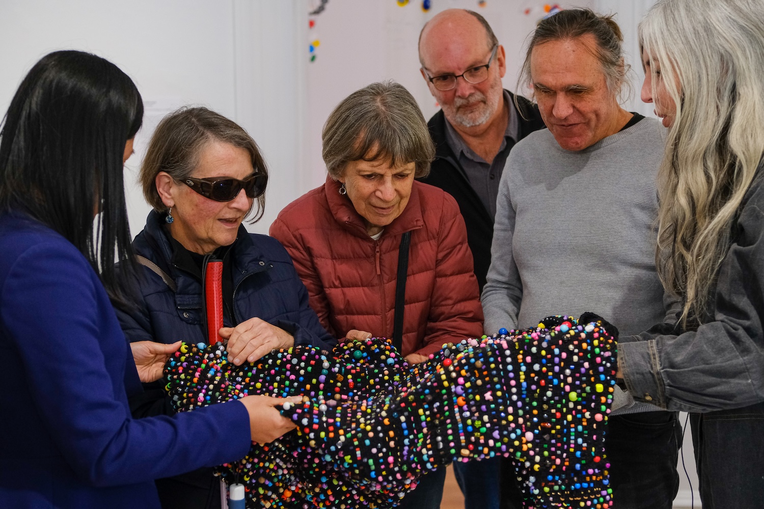 A group of six people is gathered closely, engaging with a tactile art piece during a gallery tour. The participants have varied expressions of interest and curiosity. The artwork is a textured fabric covered in numerous small, colorful beads. The beads are bright and multicolored, creating a vibrant surface. Two individuals are holding the fabric, allowing others to touch and feel the texture.