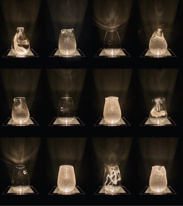 Twelve glass objects are illuminated from underneath casting yellow shadows across a black wall.