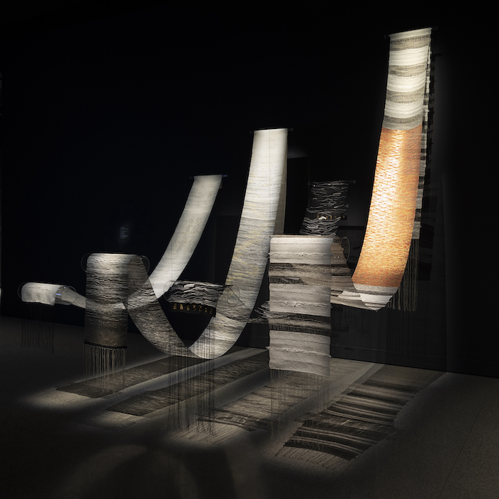 Four long paper artworks hang gracefully in a dark room.