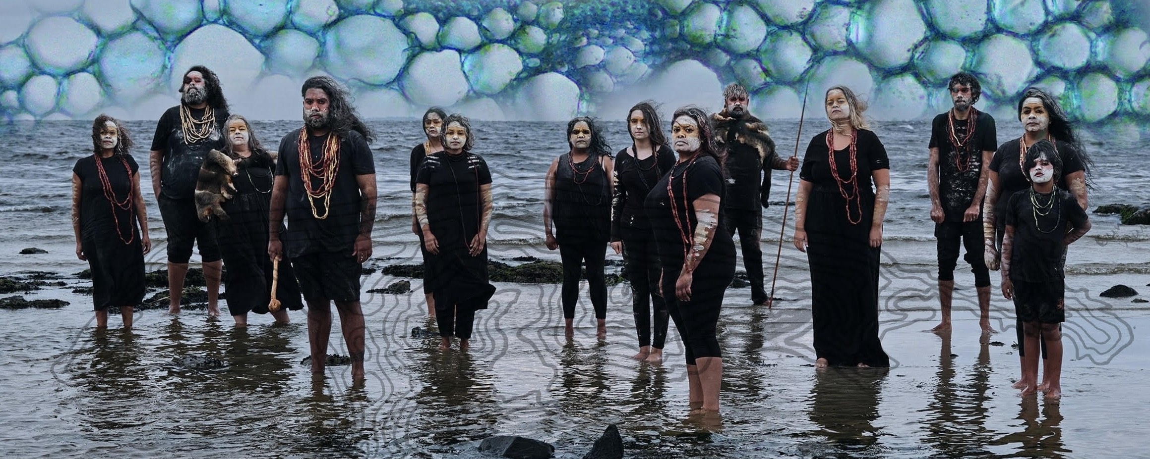 A group of people with white painted patterns, standing in ocean water with a cell-like graphic above them.